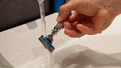 Tête de rasoir rincée sous le filet d'eau du robinet, au-dessus d'un lavabo blanc