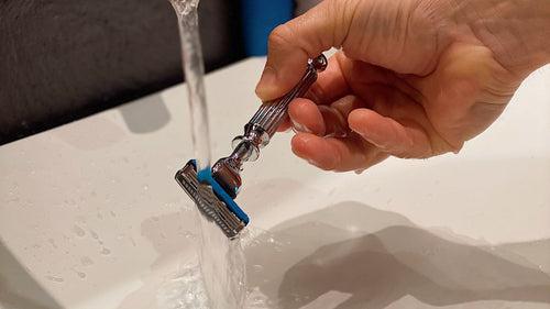 Tête de rasoir rincée sous le filet d'eau du robinet, au-dessus d'un lavabo blanc