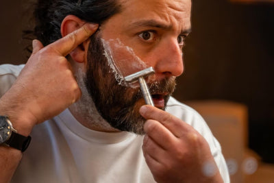 Homme se rasant la joue au rasoir de sûreté en métal, la peau tendue du bout des doigts, mousse de rasage sur la barbe