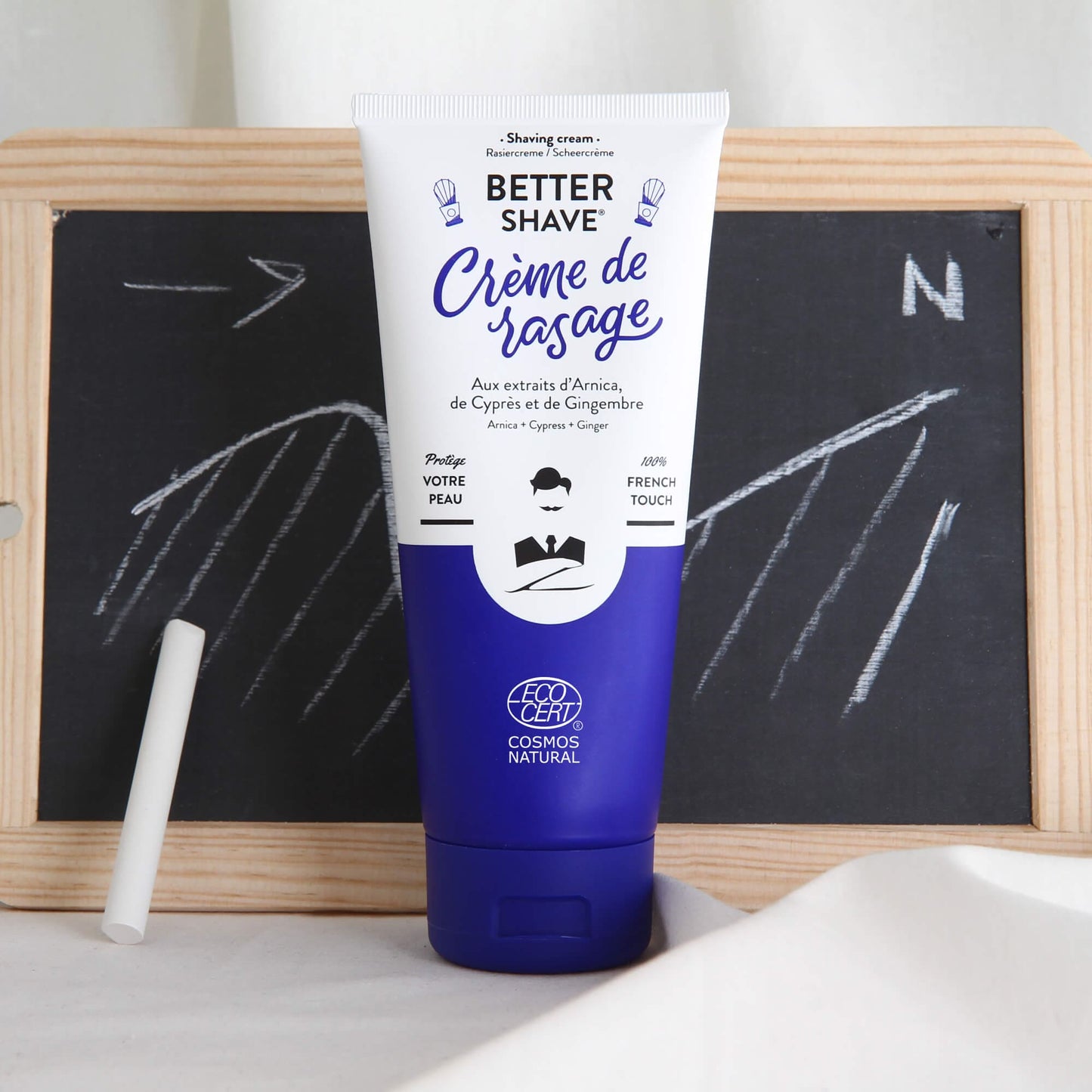 Crème de rasage Monsieur BARBIER BETTER SHAVE devant un tableau noir avec des craies, la boussole tracée suggérant la précision et l'orientation claire du produit, emballage bleu vif avec des éléments graphiques blancs.