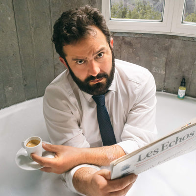 Le fondateur de Monsieur BARBIER assis dans une baignoire, en chemise et cravate, lisant le journal "Les Échos" et tenant une tasse de café, avec un produit de la marque en arrière-plan.