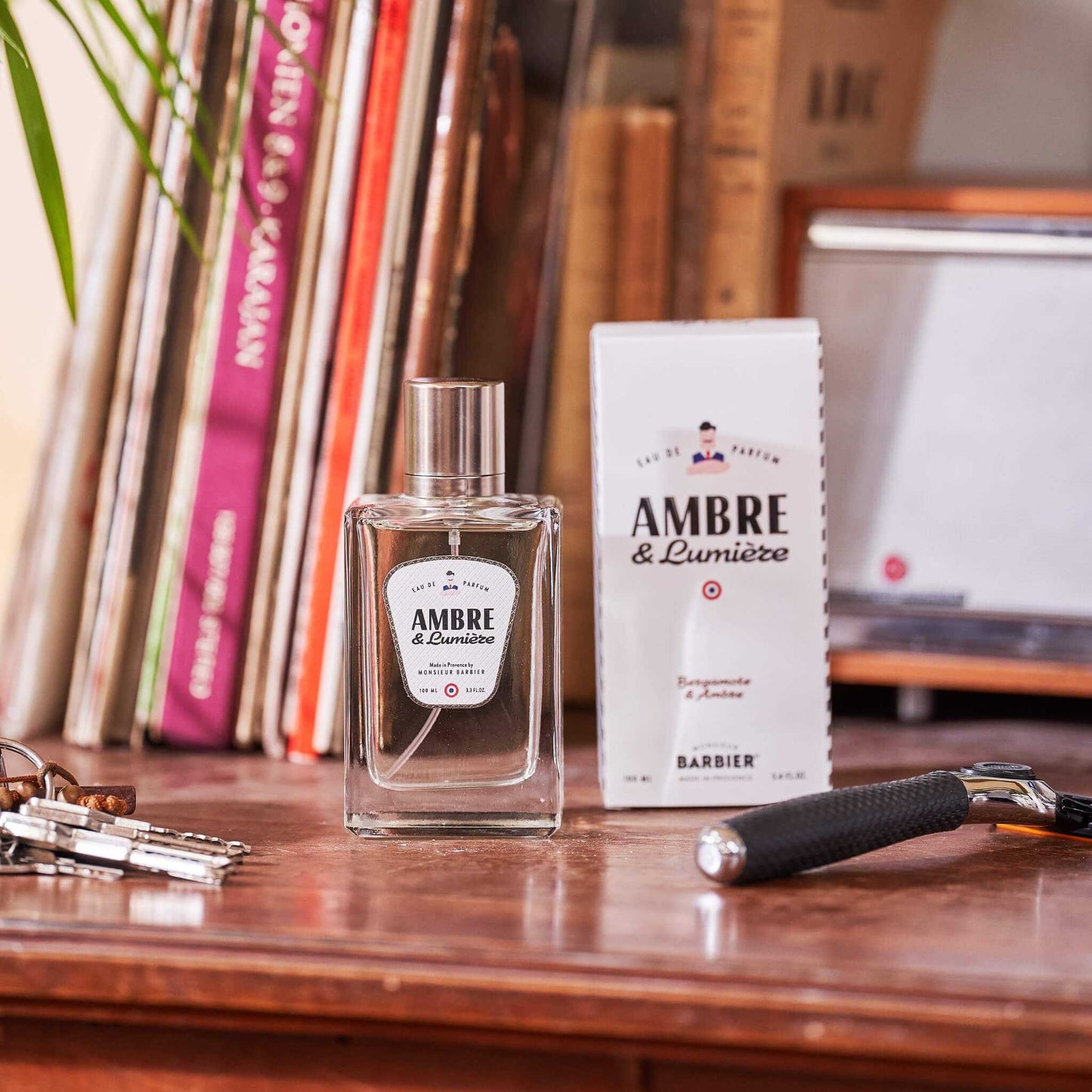 Flacon de parfum Monsieur BARBIER AMBRE & Lumière 100 ml posé près de son étui blanc sur un bureau en bois, notes de bergamote et ambre, ambiance vintage avec livres et rasoir.