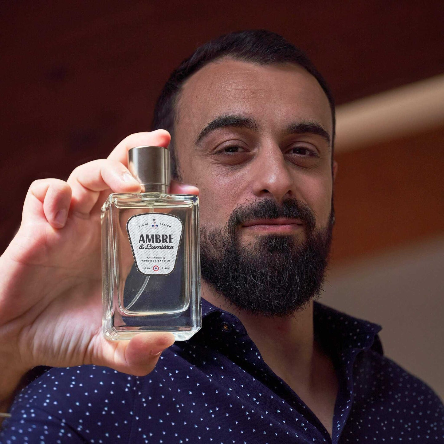 Homme barbu tenant un flacon de parfum Monsieur BARBIER AMBRE & Lumière 100 ml, étiquette blanche et noire, ambiance chaleureuse en intérieur boisé.