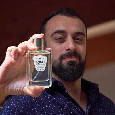 Homme barbu tenant un flacon de parfum Monsieur BARBIER AMBRE & Lumière 100 ml, étiquette blanche et noire, ambiance chaleureuse en intérieur boisé.
