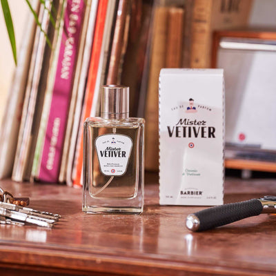 Flacon de parfum Monsieur BARBIER MISTER VETIVER 100 ml posé près de son étui blanc sur un bureau en bois, notes ozoniques et vétiver, ambiance vintage avec livres et rasoir.