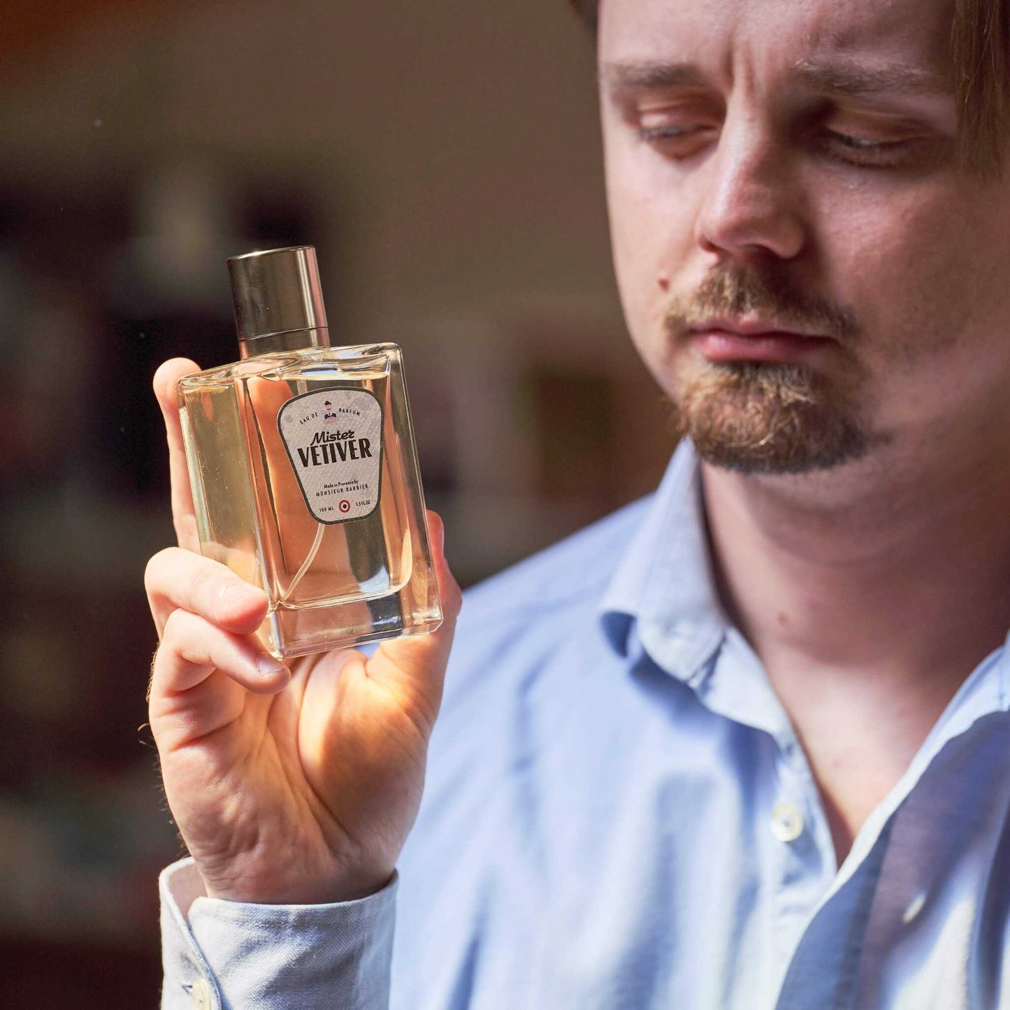 Homme moustachu en chemise bleue contemplant un flacon de parfum Monsieur BARBIER MISTER VETIVER 100 ml, étiquette blanche et noire, lumière naturelle chaleureuse.