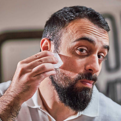 Homme barbu appliquant une pierre d'Alun Monsieur BARBIER sur sa joue après le rasage, geste apaisant et naturel, ambiance lumineuse en intérieur.